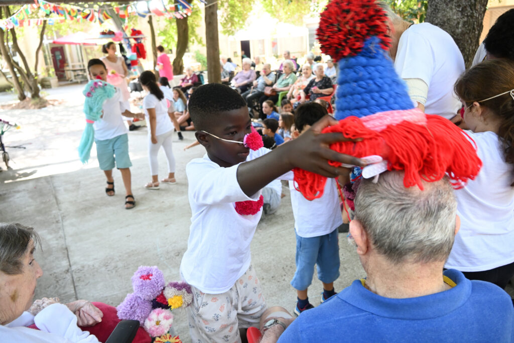 un enfant portant un nez rouge met un chapeau fait de pompons et de laine à une homme âgé assie.