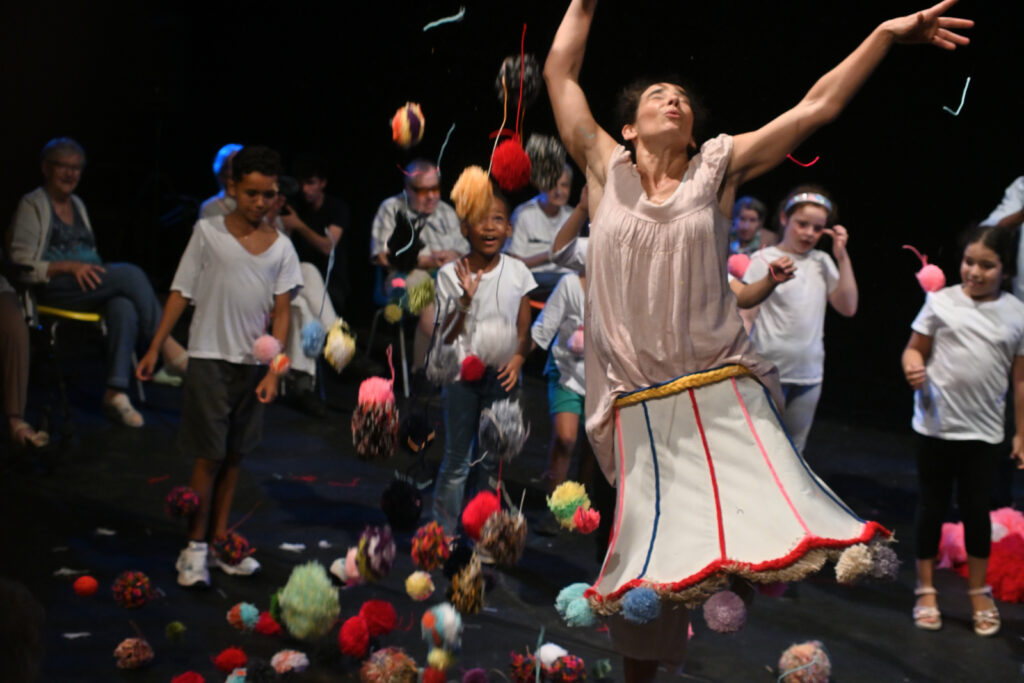 pluie de pompons sur une danseuse avec une robe en abat jour et des enfants et des personnes âgées avec des hauts blancs en arrière plan.